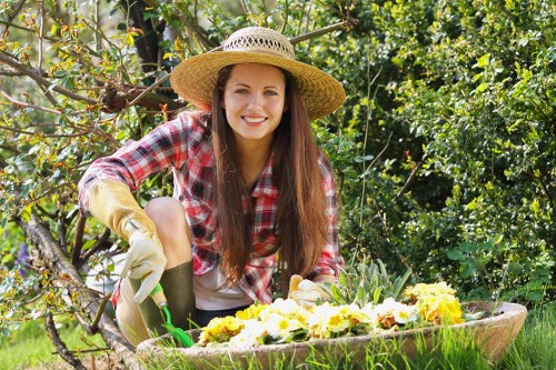 A young woman with long brown hair, wearing a straw sunhat, a red and white checkered shirt, and gardening gloves, is kneeling in a lush garden. She is smiling while tending to a flower bed filled with blooming yellow and white flowers, possibly pansies or violas. Behind her, vibrant green foliage and trees create a dense natural backdrop, with sunlight filtering through the leaves. The garden features a well-maintained lawn with dense grass surrounding the flower bed, and a mature tree branch is visible in the foreground, partially framing the scene. The environment appears vibrant and healthy, indicative of regular garden maintenance which a professional gardening service like Hedge Trimming Barkingside could support. The overall setting suggests a pleasant, outdoor landscaped space typical of residential gardens in Barkingside, Essex, with natural textures and colours contributing to a welcoming and well-kept appearance.