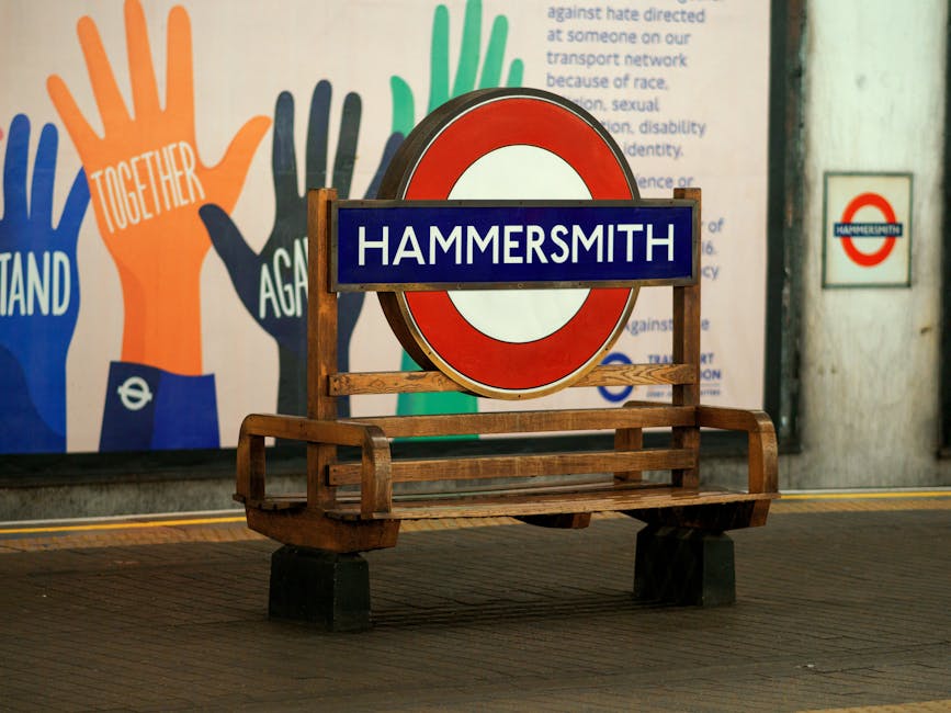 A wooden garden bench with black supports positioned in front of a large circular Hammersmith London Underground station sign on a wall. Behind the bench, there is a colorful mural featuring stylized
