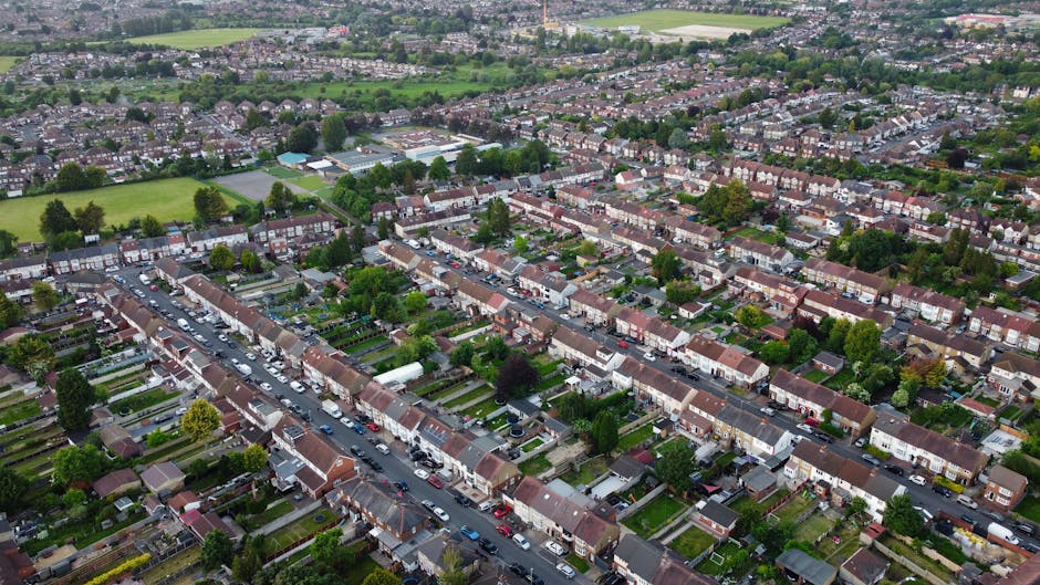 An aerial view of a residential neighbourhood showing a row of terraced houses with small front gardens and parked cars along the street. Behind the houses, there are numerous backyards with well-main